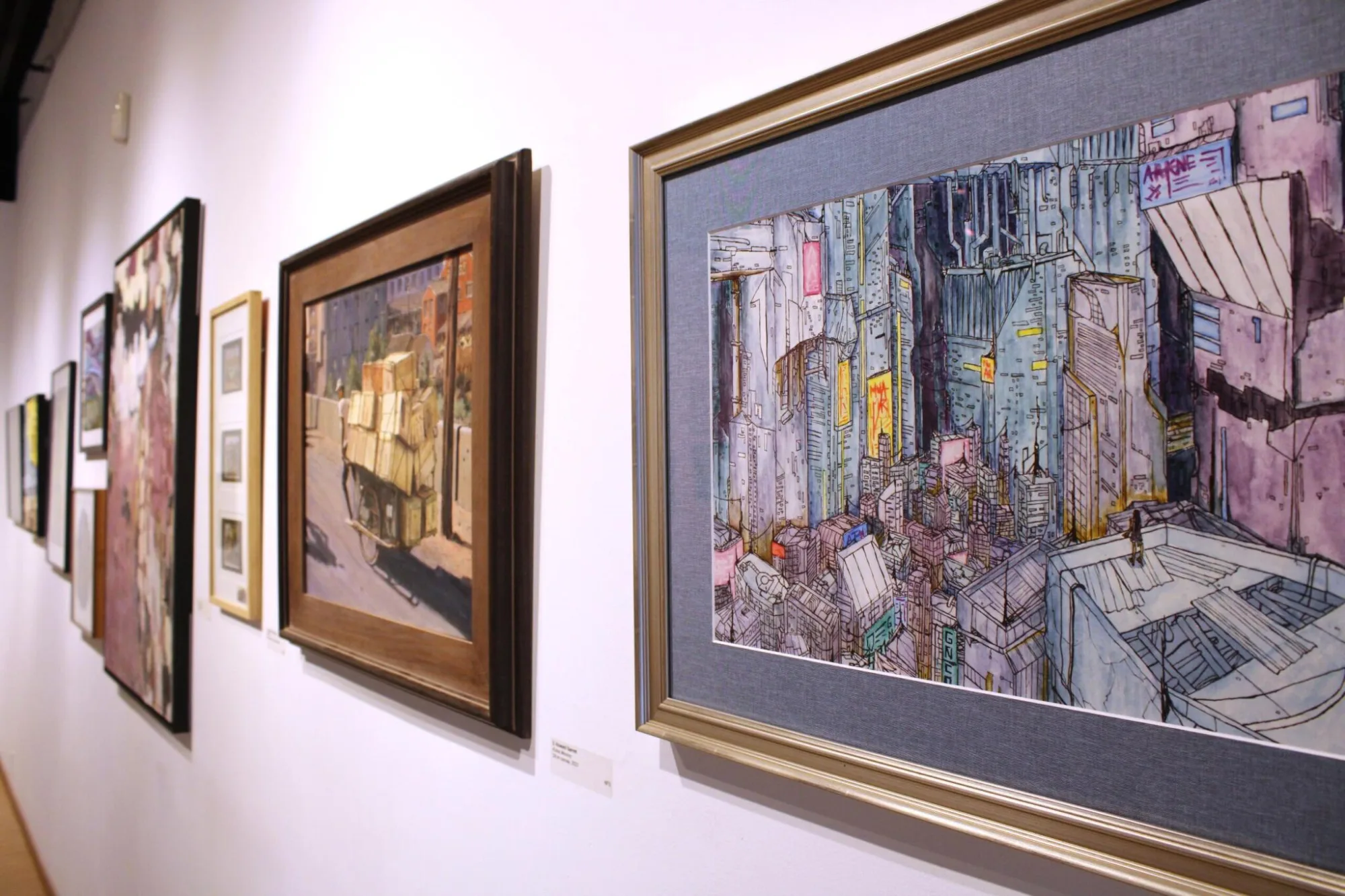 Art gallery wall displaying framed paintings, including urban and rural scenes, in a variety of styles and colors.