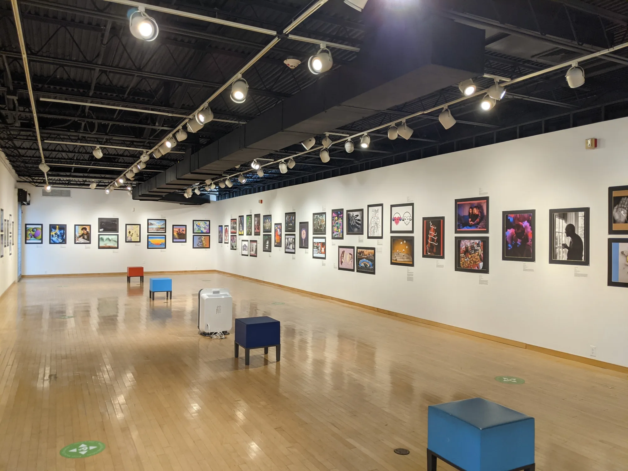 Empty indoor art gallery with framed artwork hung along the walls, with directional track lighting, and light colored hardwood flooring.