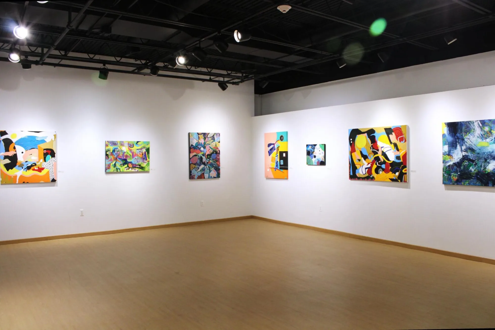 An art gallery with abstract paintings on white walls, wood flooring, and overhead lighting.