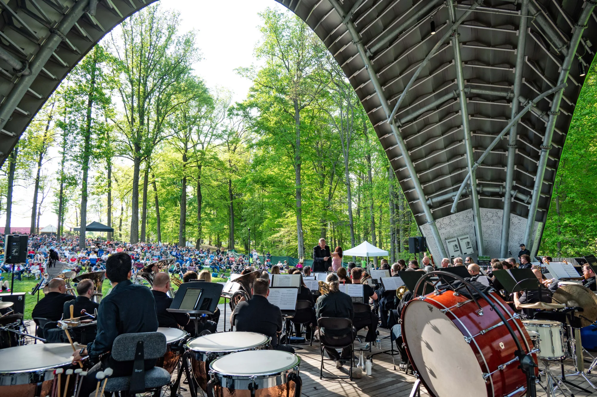 Orchestra performing on an outdoor stage in a forested area, with a large audience seated on the grass in the background.