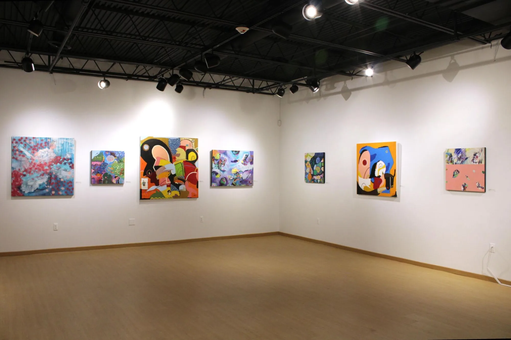 An art gallery with abstract paintings on white walls, wood flooring, and overhead lighting.