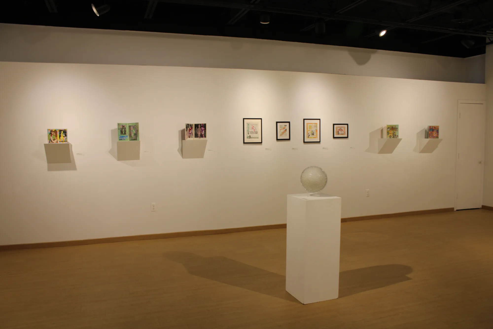 Sphere sat on white podium in the center of the light hardwood floor, behind is a wall with artwork installed along the length.