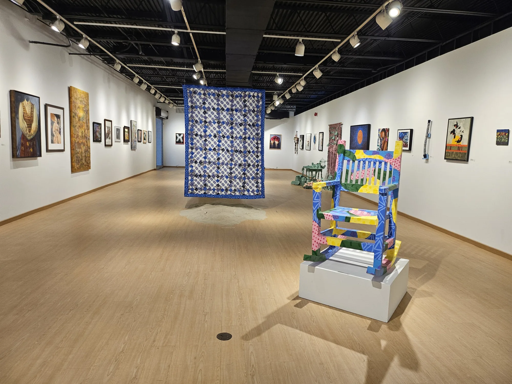 Artwork hangs at eye level along the walls, a patterned quilt hangs down into the middle of the gallery, a colorful painted wooden rocking chair placed in the front right corner, directional track lighting line the ceilings, and light colored hardwood flooring.