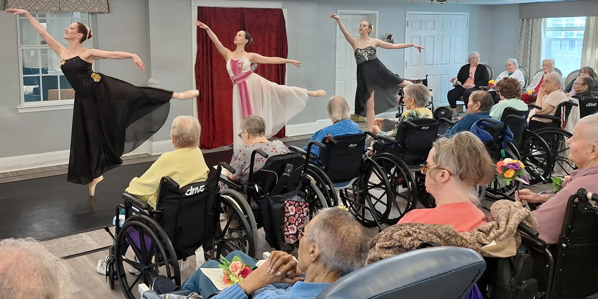 Three ballerinas in the arabesque stance performing for an audience of elderly women sitting in wheelchairs holding flowers wearing flowy gowns.