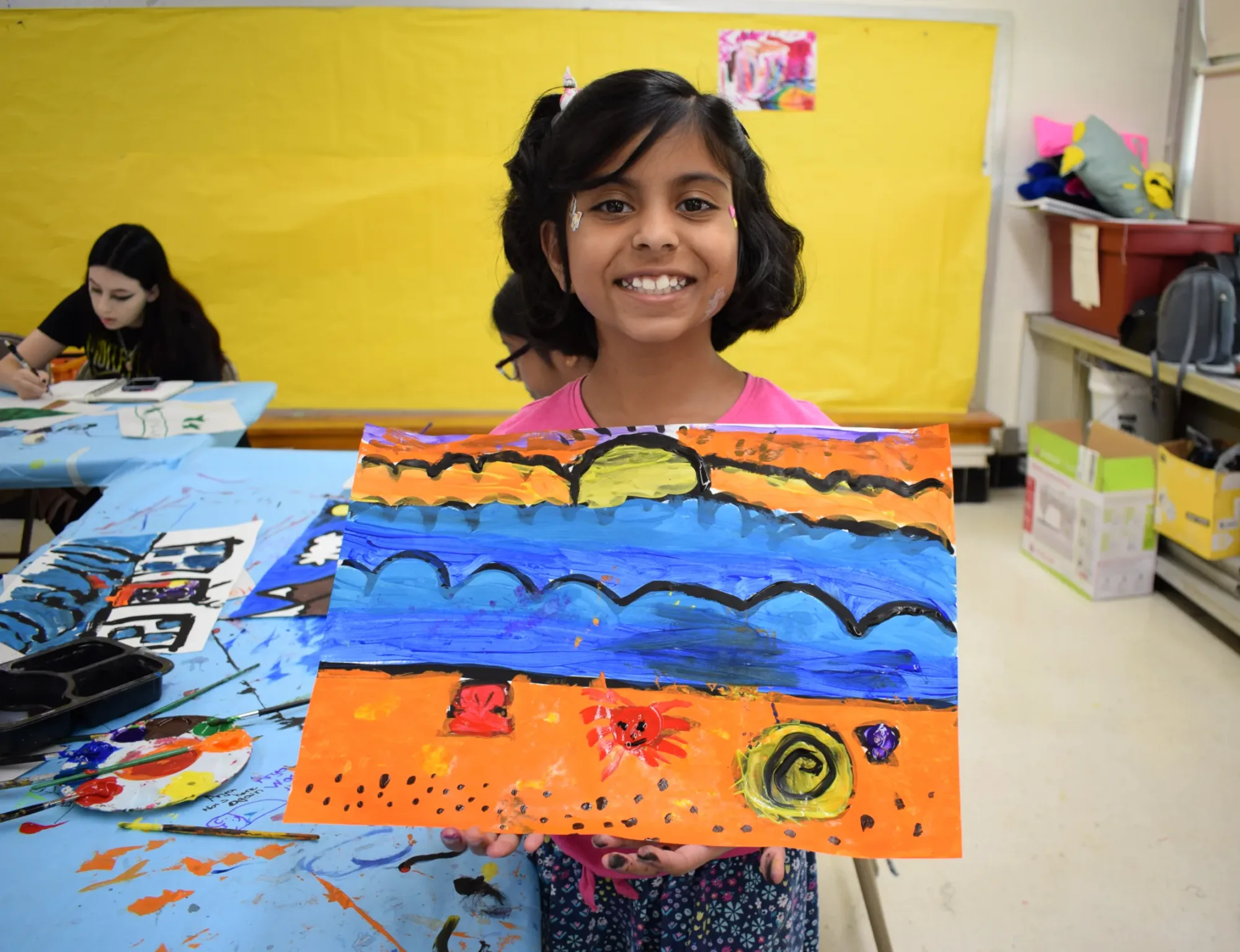 Girl smiles and shows her painting of sun over ocean.