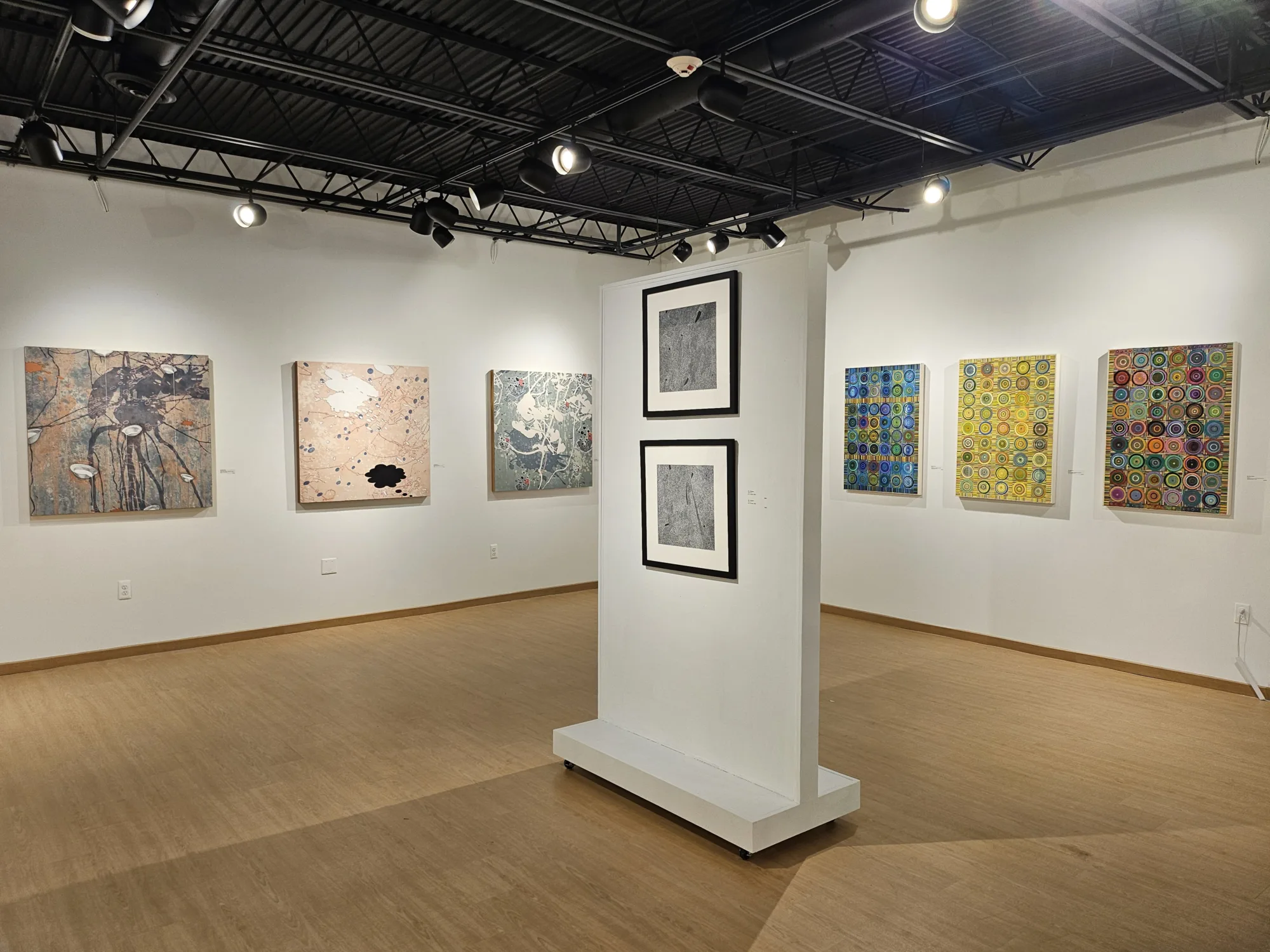Artwork hangs along two indoor gallery walls that meet at corner, diagonal from the corner sits a rollable makeshift wall with two framed pieces of artwork hanging horizontally. Directional track lighting above and light hardwood flooring.