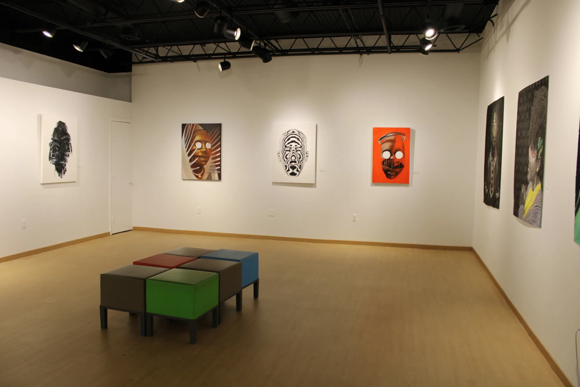 Art gallery with abstract mask paintings on white walls, colorful stools in the center, and overhead lighting.