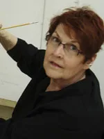 Woman with glasses and short red hair stands sideways looking forward with a art tool in her hand.