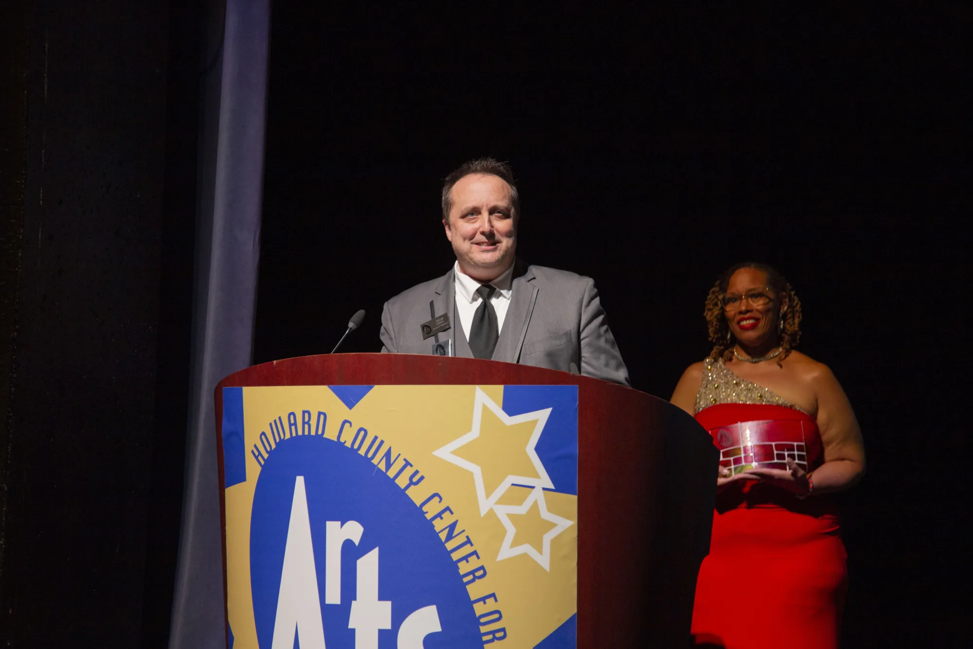Smiling man in suit stands at podium and a women in red stands behind him.