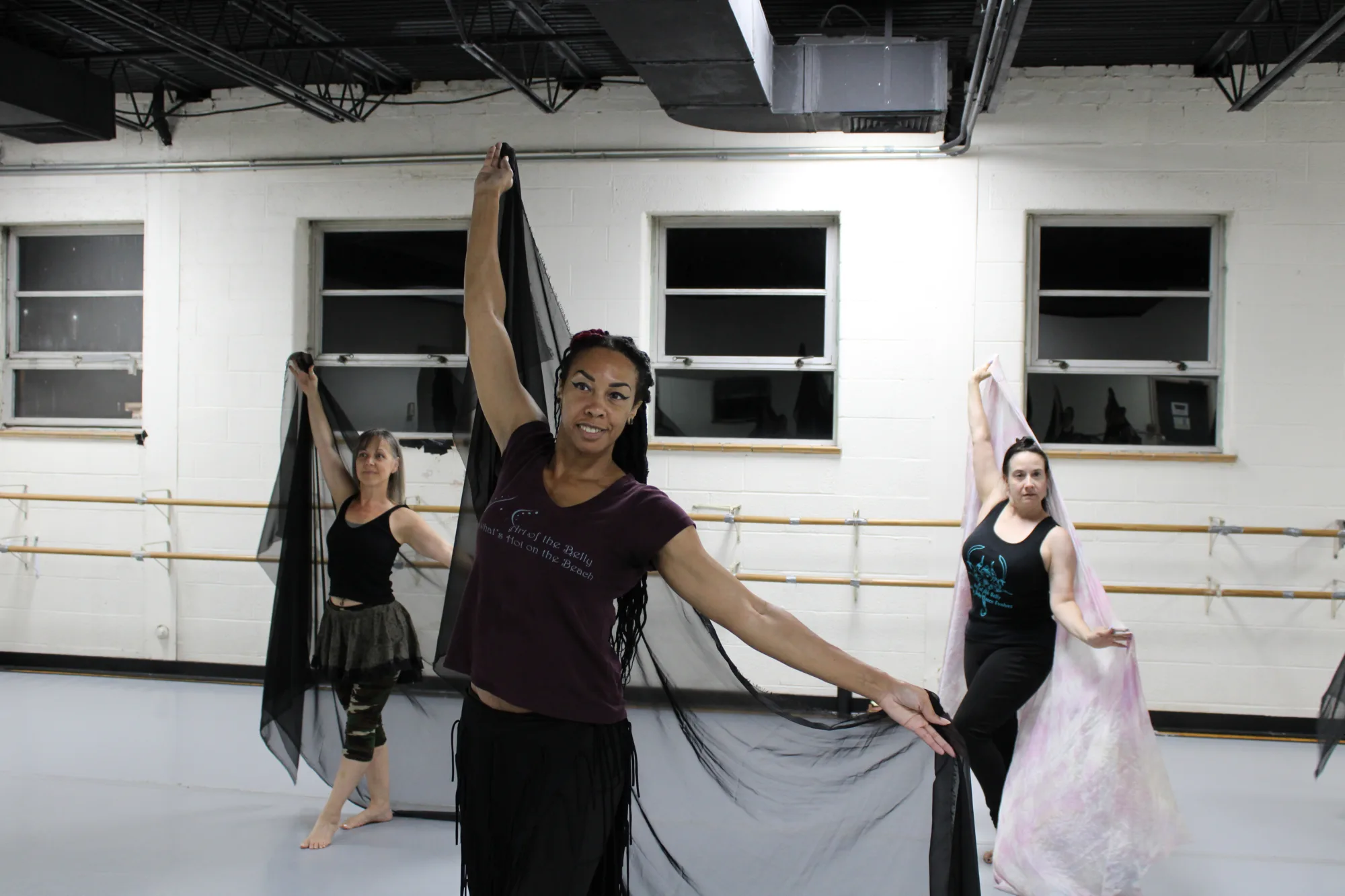 Three women hold scarves at an angle behind themselves in a dance studio