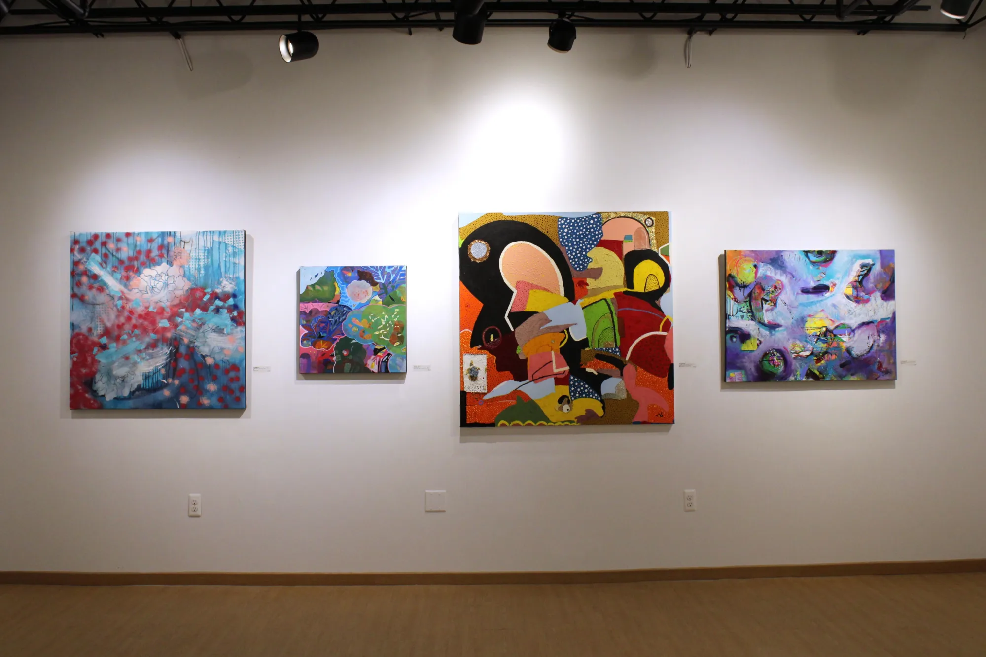 Four vibrant abstract paintings hang on a gallery wall, each featuring a mix of colors, shapes, and patterns under overhead lighting.