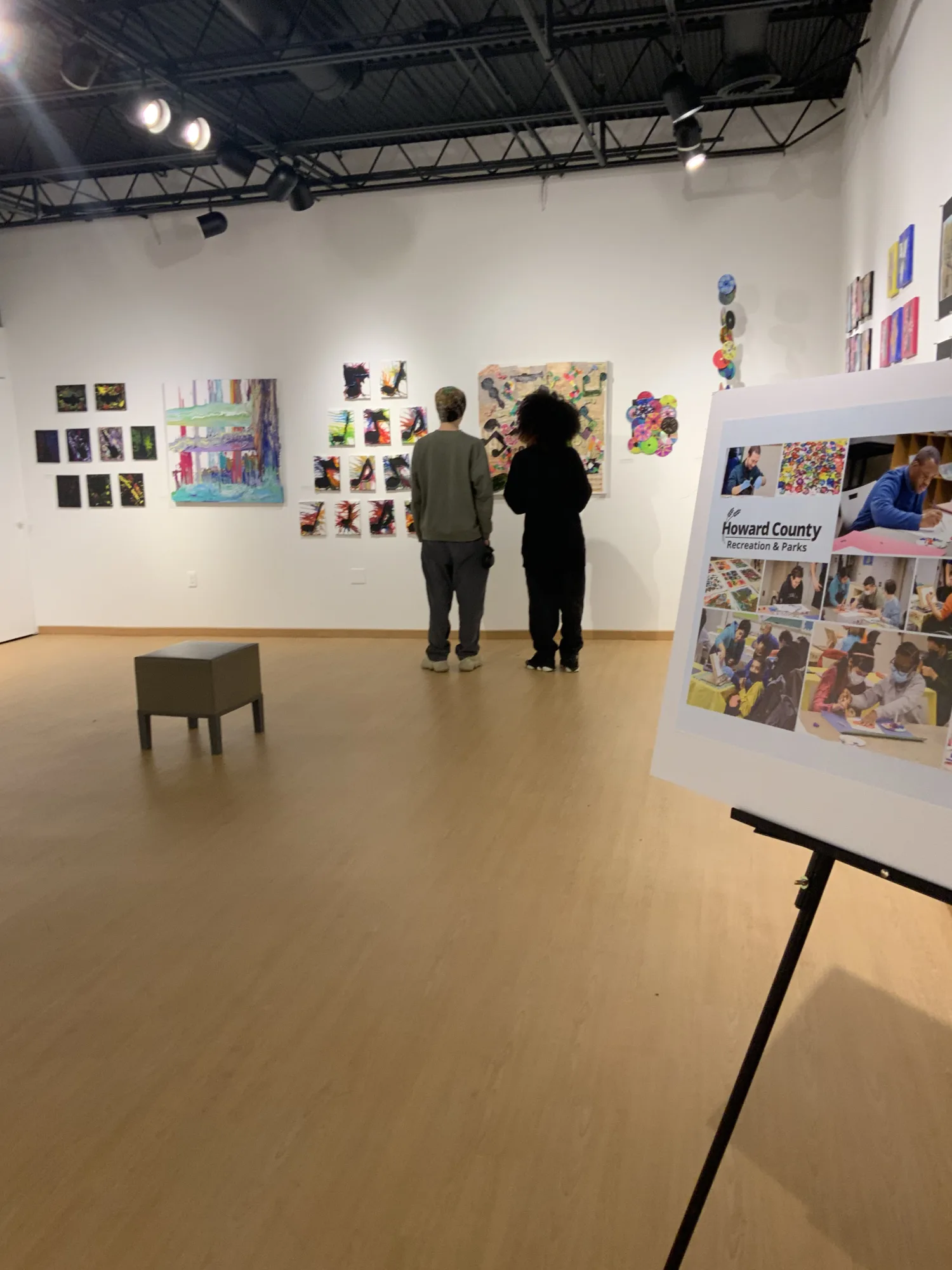 Two people face a wall with collections of artwork hung, additional artwork hung on right wall, with a single stool placed in the middle of the floor.