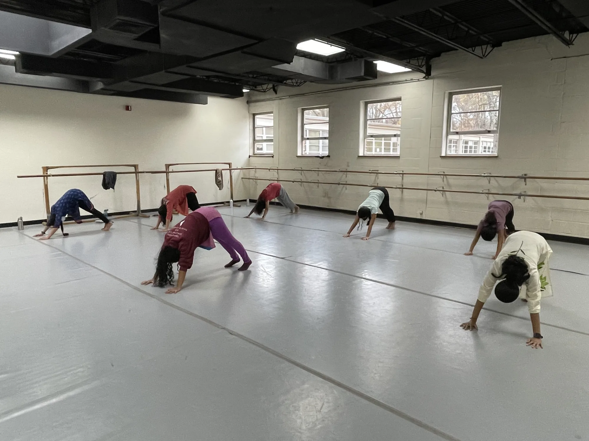 A group of adults doing yoga in downward facing dog pose in a dance studio with windows behind them
