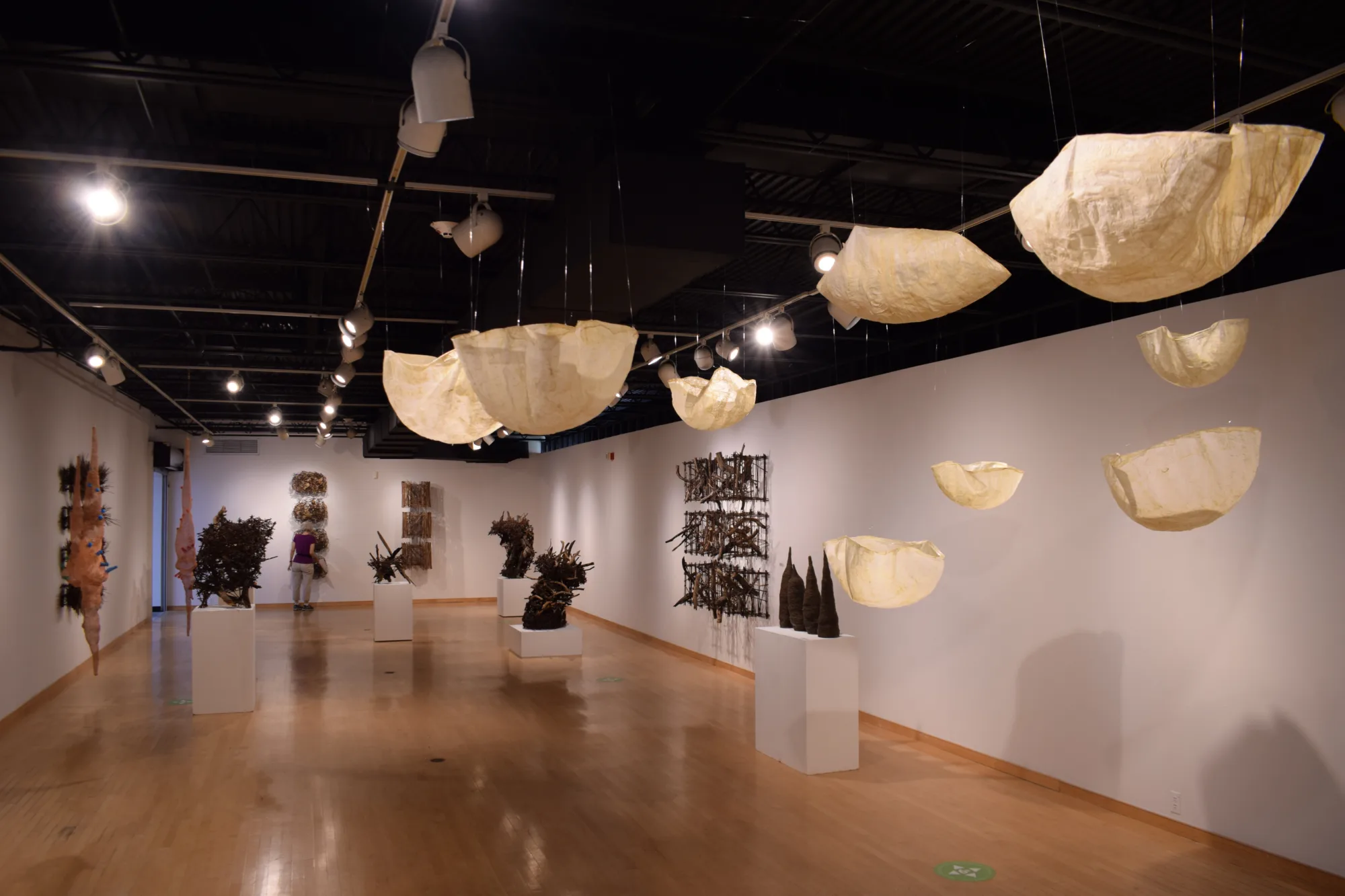 Art gallery with hanging fabric sculptures, wall-mounted artworks, and various sculptures on pedestals. The room is lit by ceiling spotlights.