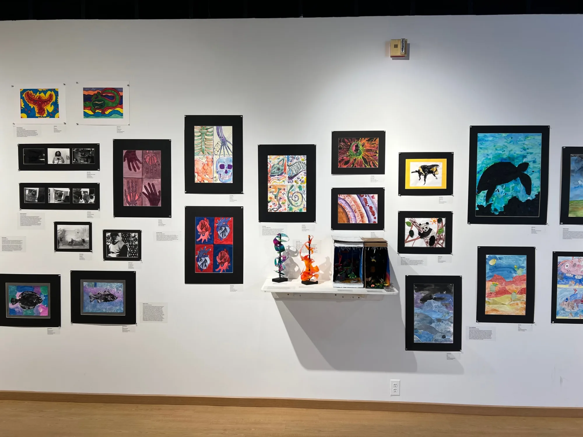 Art gallery with 2-D student artwork hung salon-style with all black mats and shelf with 4 small sculptures.