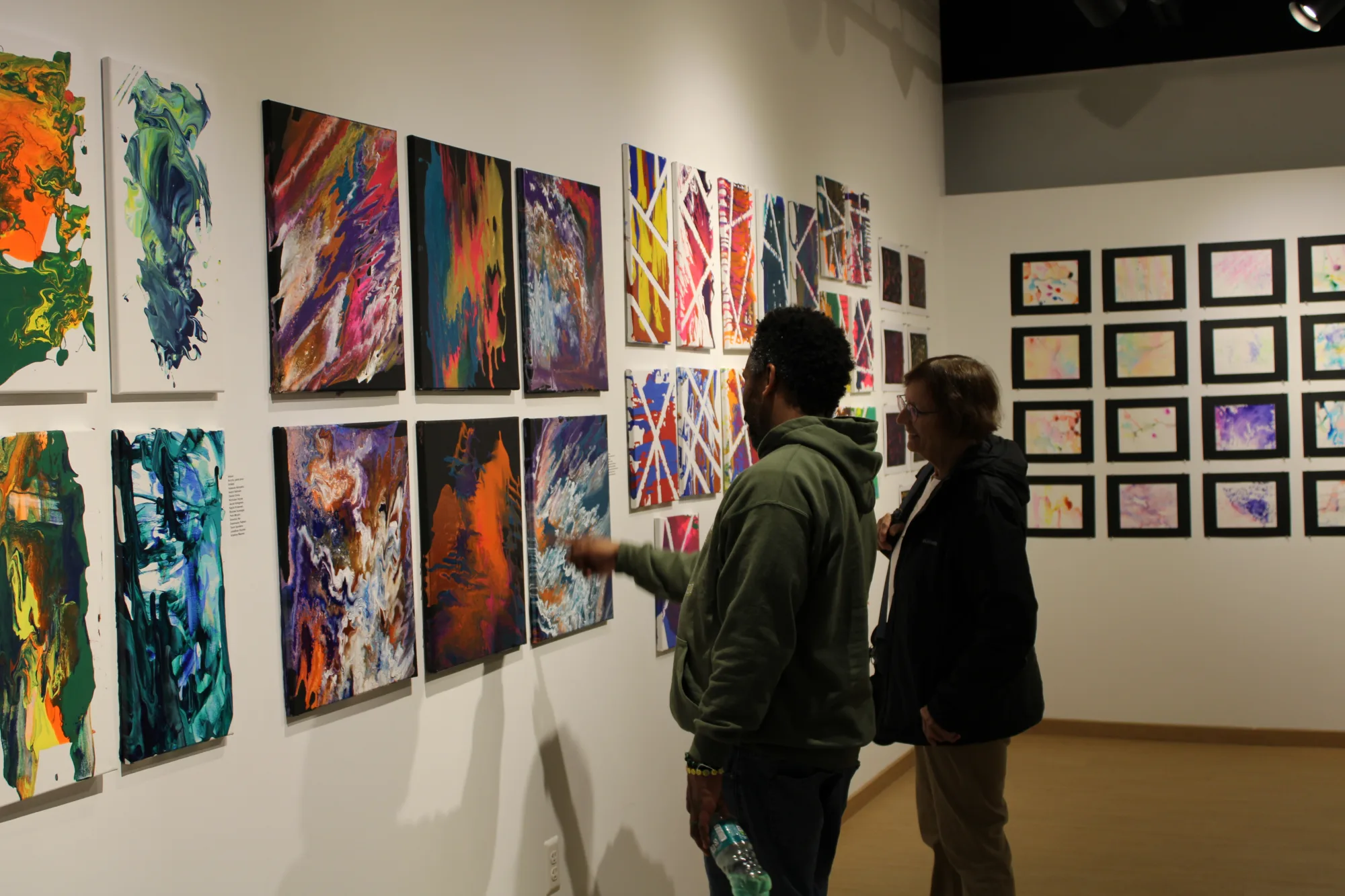 Artwork hangs along two indoor gallery walls that meet at a corner, with two visitors standing up close looking at the left wall.