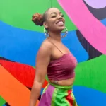 Woman smiles standing in front of colorful abstract mural wearing a pink top and multicolored skirt.
