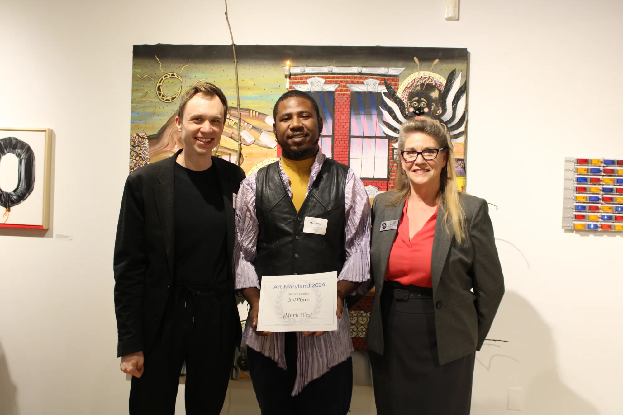 Two men, one holding a certificate, and one woman, stands together in front of a painting on a art gallery wall.