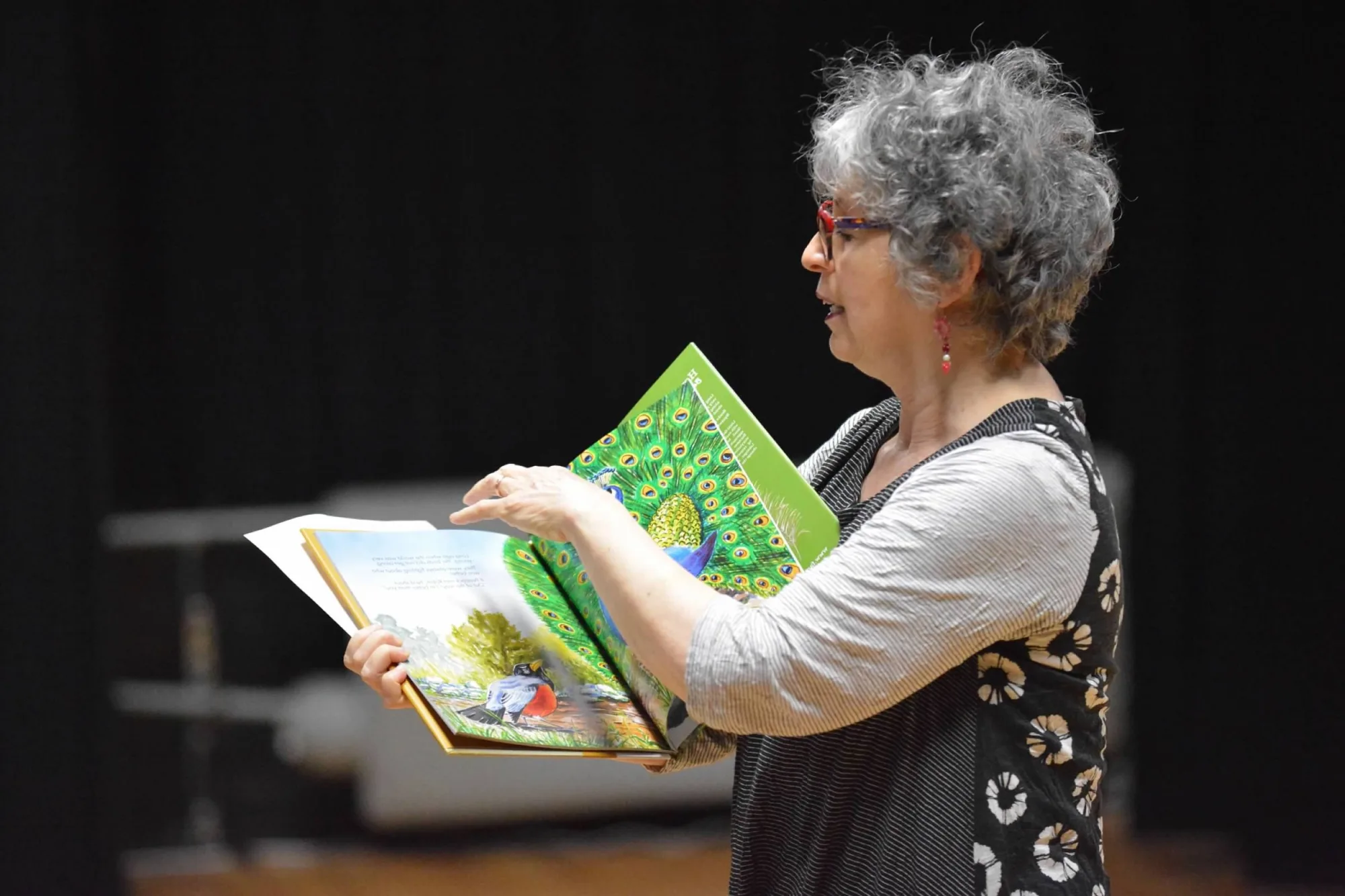 Woman stands holding open a book with birds illustrated on the pages, reading in a black and white top.