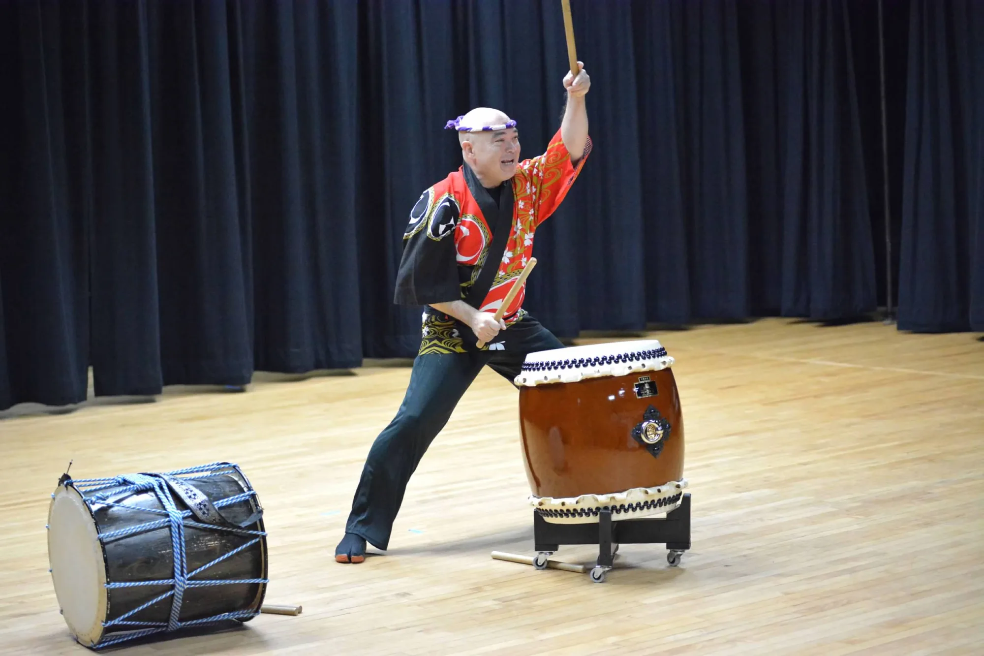 Asian man on a stage standing behind a drum with one arm raised. Another drum is pictured on the floor next to him.